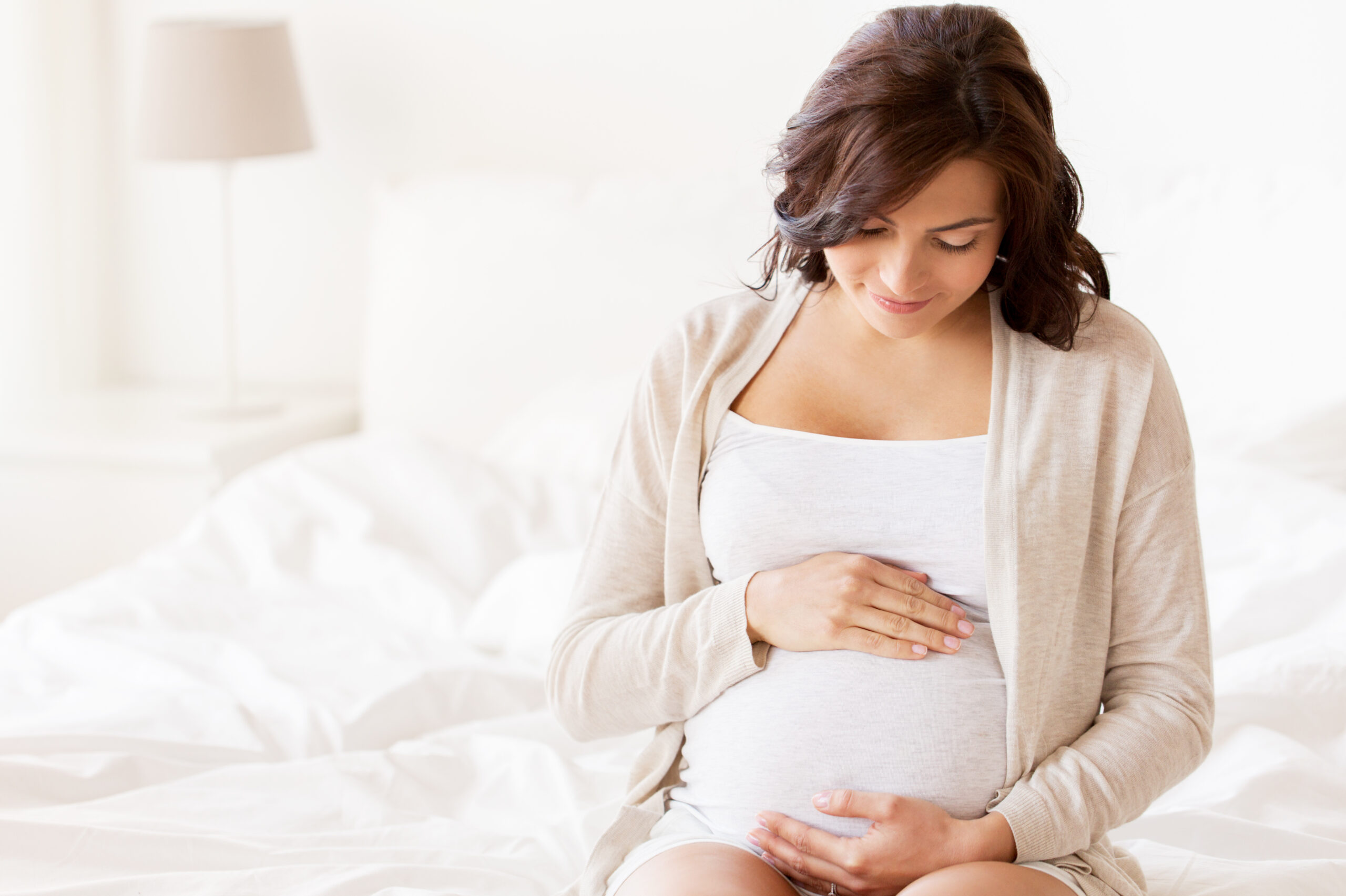 pregnancy, rest, people and expectation concept - happy pregnant woman sitting on bed and touching her belly at home